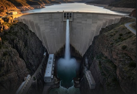 An aerial view of a massive hydroelectric dam. A large concrete wall spans a wide canyon, holding back a huge lake. At the base of the dam, a power station is visible, with water churning below. Steep, forested hillsides surround the dam and the water.